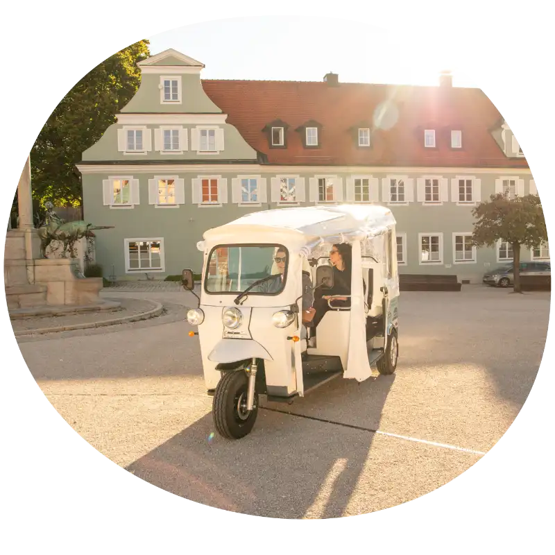 Foto eines E-Tuk-Tuks von Tuk Rum in Kempten (Allgäu) mit einer Fahrerin und einer Passagierin bei schönem Wetter.